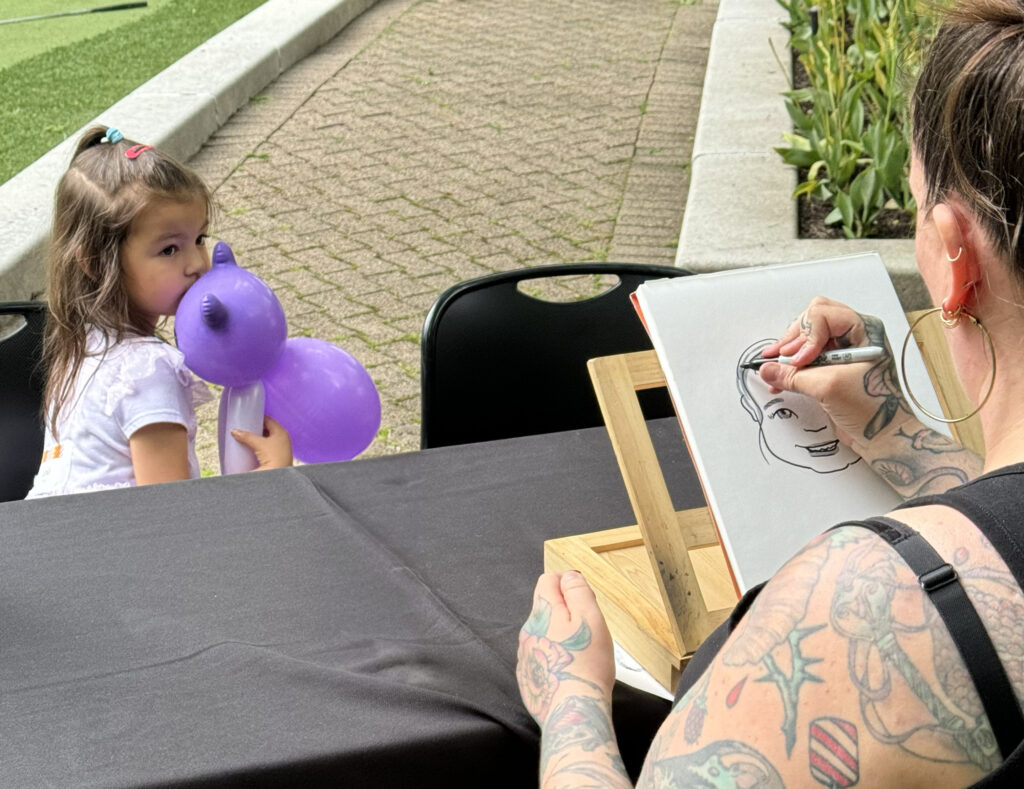 A caricature artist drawing a small girl. You can see behind the caricature artist's head to both the drawing and the girl, who is holding a pink cat balloon.