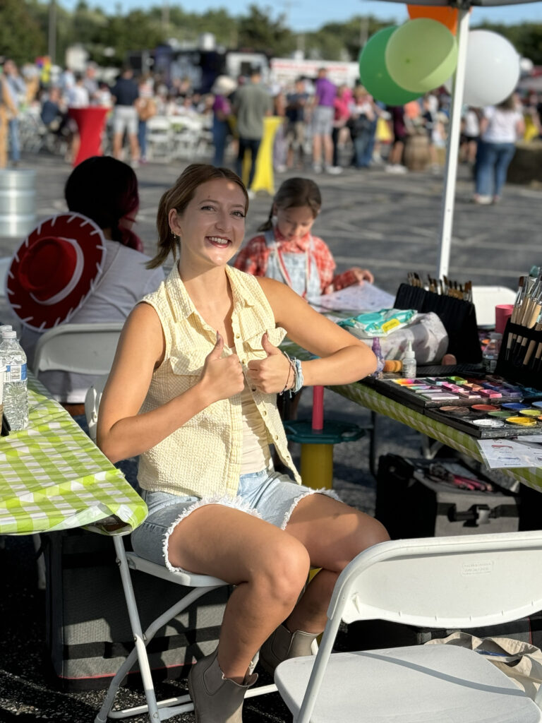 Face painter and costume character Olivia seated under a tent and giving the thumbs up at a large outdoor event