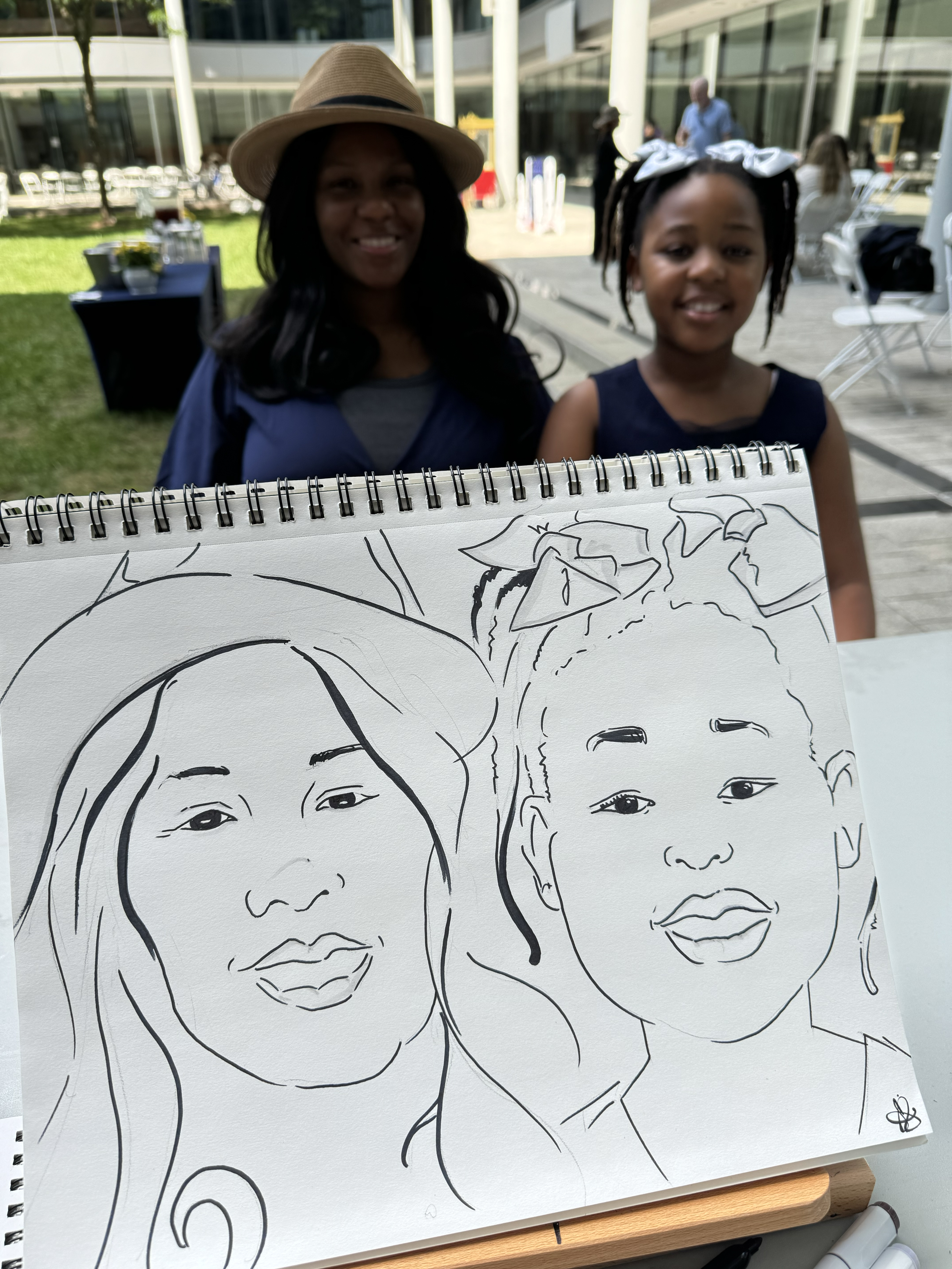 A mother and daughter sitting to have their caricatures drawn in NH. The POV is by the caricature artist, and you can see both the family and the drawing in the photo.